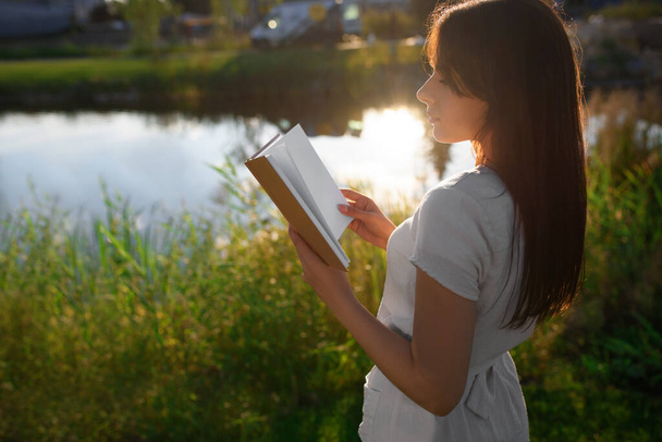 Young woman reading book near lake on sunny day - Фото, зображення