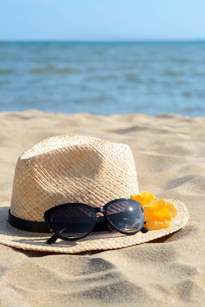 Hat with beautiful sunglasses and flowers on sand near sea. Space for text - Fotoğraf, Görsel