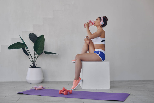 Fit young woman in sport clothing drinking water while relaxing after training indoors - Photo, Image