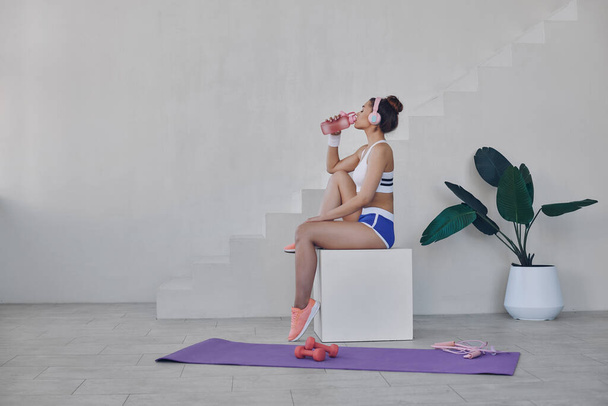 Beautiful young woman in sport clothing drinking water while relaxing after training indoors - Foto, Imagem