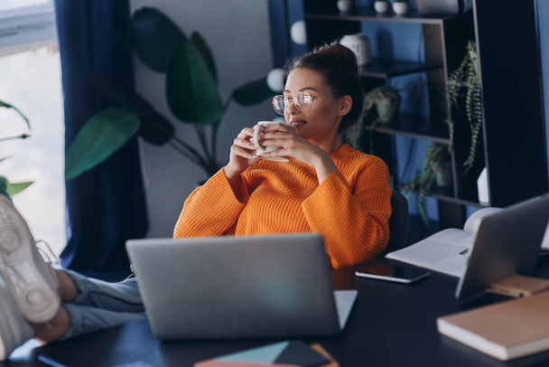 Beautiful woman holding her feet on desk and enjoying coffee while sitting at her working place in office - Foto, afbeelding