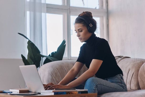 Confident young woman in headphones using laptop while sitting on the couch at home - Fotografie, Obrázek