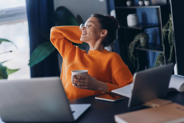 Happy young woman holding coffee cup and keeping her feet on desk while sitting in office - Foto, afbeelding