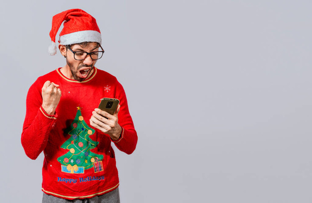 Excited guy in christmas hat looking at cellphone and celebrating. People in santa hat looking at cell phone excited, Excited young man in christmas hat using cellphone celebrating - Foto, imagen