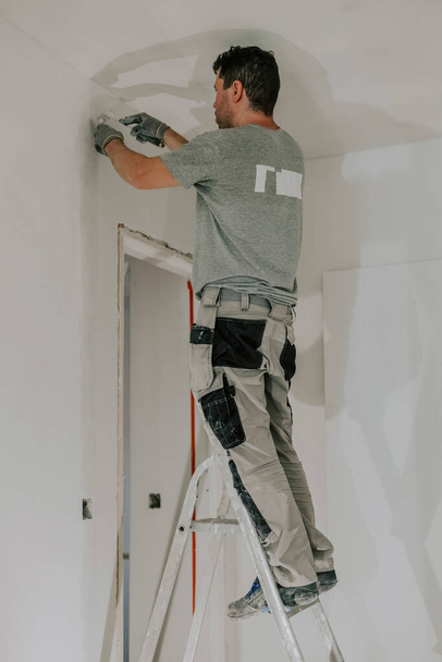 A handsome young Caucasian man in a gray uniform and gloves standing on a stepladder puttying two small spatulas on the wall in the doorway in the evening with a building lantern on, close-up side view. Construction concept. - Foto, Imagem