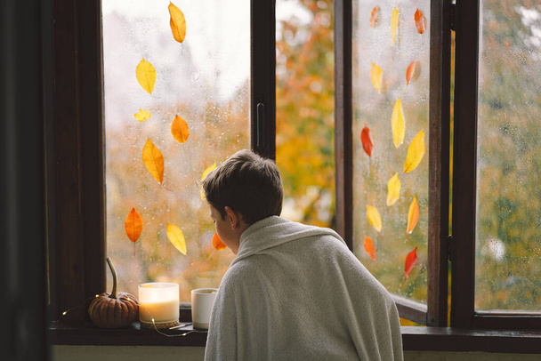 Ένα χαριτωμένο αγόρι τυλιγμένο σε μια κουβέρτα κοιτάζει έξω από το ανοιχτό παράθυρο στην υπέροχη φθινοπωρινή φύση. Φθινοπωρινή διακόσμηση. Ζεστή φθινοπωρινή διάθεση. Ευχαριστίες. Απόκριες. - Φωτογραφία, εικόνα