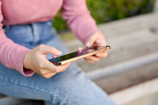Zrelaksowana młoda Azjatka w luźnych ciuchach, używająca smartfona do oglądania wideo lub grania w gry mobilne siedząc na ławce w parku. przycięte i zbliżenie obrazu - Zdjęcie, obraz