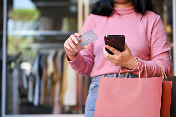 cropped shot, Young Asian woman with her shopping bags holding a credit card and her smartphone, standing in front of clothes store. - Foto, Bild