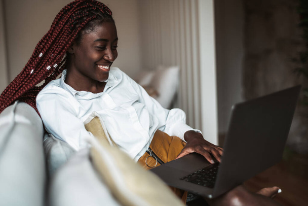 Portrait d'une jeune femme africaine heureuse et indépendante avec de longues tresses, assise sur le canapé d'un salon et utilisant l'ordinateur portable pour parler à son amie par appel vidéo ; une femme noire avec un netbook - Photo, image
