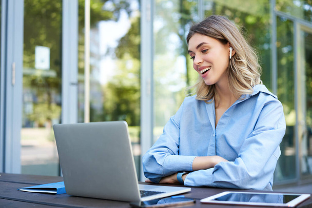 Portrait of corporate woman working outside office on remote, sitting with laptop, wireless earphones, attend online video conference. - Foto, afbeelding