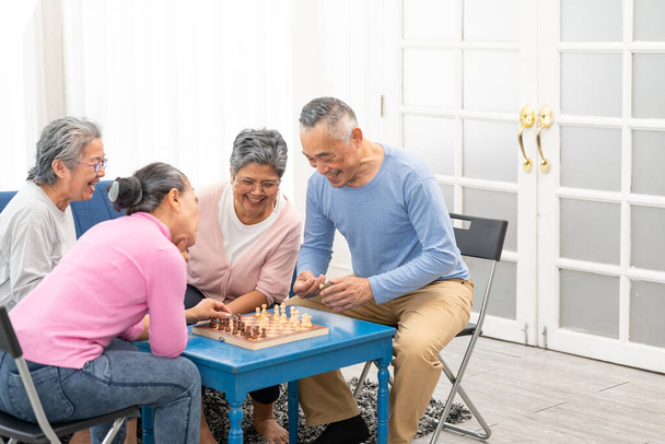 Ασιάτες ηλικιωμένοι άνδρες και γυναίκες σε casual παιχνίδι σκάκι στο γηροκομείο, κάθονται γύρω από το τραπέζι. Ασιάτης πρεσβύτερος άνδρας περνάει ελεύθερο χρόνο. Χαρούμενο χαμόγελο Μεγαλύτερος ώριμος άνθρωπος απολαμβάνουν δραστηριότητα στο σπίτι. - Φωτογραφία, εικόνα