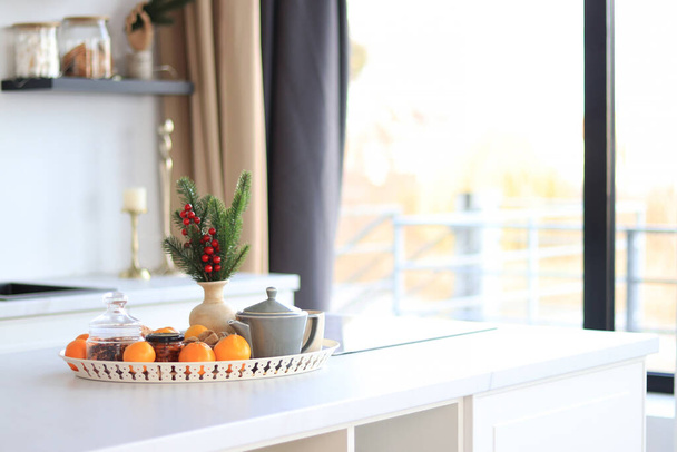 Asetelma mandariinit, teekannu ja asteikot kuusen oksa keittiön pöydällä, kirkas aurinkoinen sisustus. Juhlatunnelma keittiössä, valikoiva keskittyminen - Valokuva, kuva
