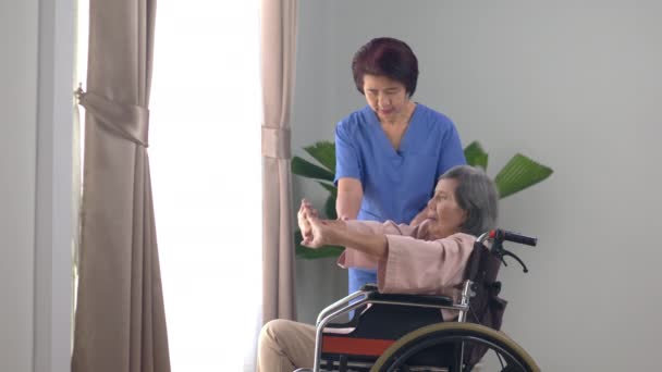 elderly asian woman doing stretching exercise on wheelchair in living room at home with physiotherapist. . - Footage, Video