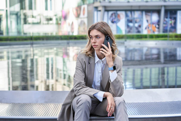 Stilvolle Geschäftsfrau im Anzug, am Brunnen vor Bürogebäuden sitzend, telefonierend. Korporative Frau wartet draußen auf jemanden. - Foto, Bild