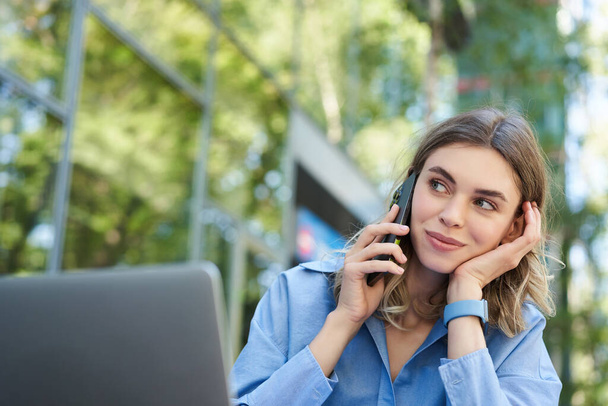 Porträt einer jungen Geschäftsfrau, die draußen im Park sitzt, mit dem Handy telefoniert und ihren Laptop benutzt. Junge Frau, Studentin, die im Freien arbeitet, telefoniert, telefoniert. - Foto, Bild