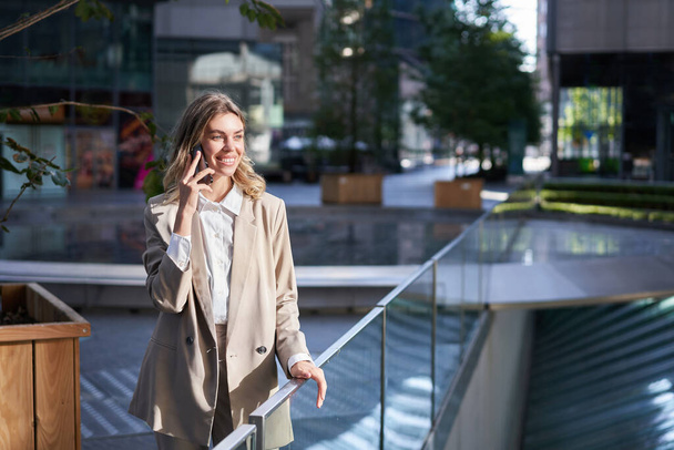 Успешная деловая женщина разговаривает по мобильному телефону с уверенной улыбкой, справляется с деловыми звонками, стоит на улице в городе. - Фото, изображение