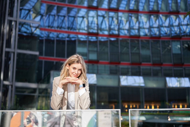 Schöne junge Frau im beigen Anzug, steht in der Nähe von Bürogebäuden im Stadtzentrum, lächelt und sieht verträumt aus. - Foto, Bild