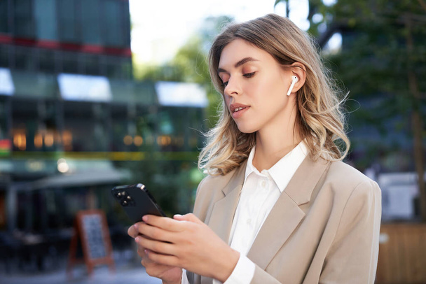Femme d'affaires prospère en costume beige, écouteurs sans fil, regardant le téléphone mobile, utilisant l'application smartphone, debout à l'extérieur dans la rue. - Photo, image