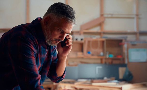 Multiracial mid adult man talking on mobile phone near woodpile in woodworking factory - Zdjęcie, obraz