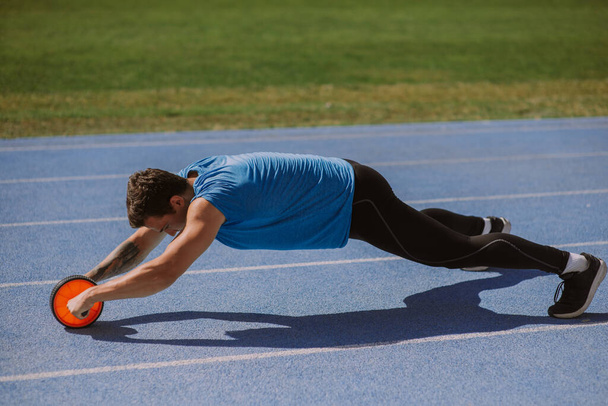 Muskularny mężczyzna ćwiczący z rolką na kółkach brzucha, na stadionie, w piękny słoneczny dzień - Zdjęcie, obraz