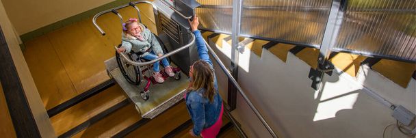 Mother with a young child living with cerebral palsy using electric wheelchair lift to access public building. Special lifting platform for wheelchair users. Disability stairs lift facility. - Photo, Image