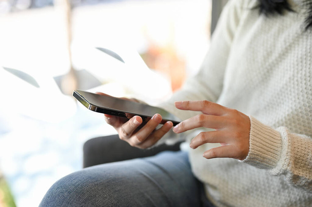 Una mujer asiática en suéter blanco usando su teléfono inteligente mientras se relaja en el café, charlando, leyendo el blog en línea, utilizando la aplicación móvil. imagen de primer plano - Foto, Imagen