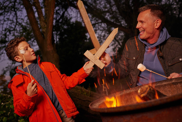 En garde, aber beobachte meinen Marshmallow. Vater und Sohn spielen mit Holzschwertern, während sie Marshmallows über dem Feuer anstoßen - Foto, Bild