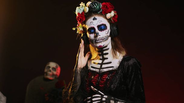 Κυρία του θανάτου ακούγοντας μουσική στα ακουστικά, φορώντας μέικ-απ κρανίου και κοστούμι Σάντα Μουέρτε τη μέρα των νεκρών. Απολαμβάνοντας διασκεδαστικό τραγούδι σε ακουστικά ήχου, γιορτάζοντας μεξικάνικη παράδοση. Χειροκίνητη βολή. - Φωτογραφία, εικόνα