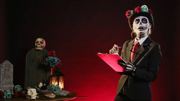 Dios de los muertos Modell, das Notizen auf Zetteln macht, sieht aus wie Weihnachtsmann mit Totenschminke auf mexikanischen Feiertagen. Todesdame Tracht mit Anzug und Hut. - Foto, Bild