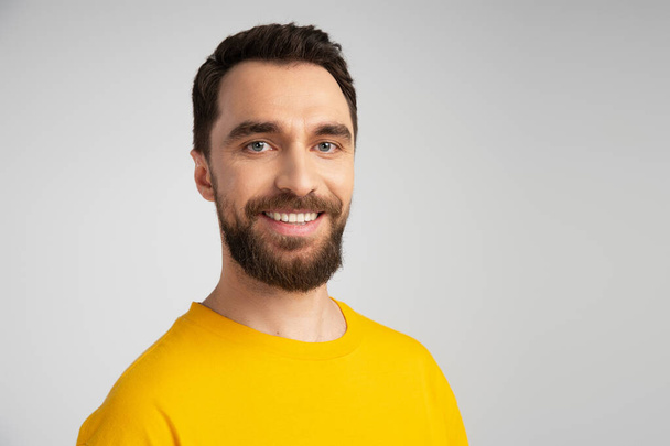 portrait of joyful bearded man in yellow t-shirt looking at camera isolated on grey - Fotó, kép
