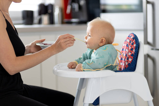 Материнская ложка кормит своего маленького мальчика в детском кресле фруктовым пюре на кухне дома. Концепция введения твердого питания. - Фото, изображение