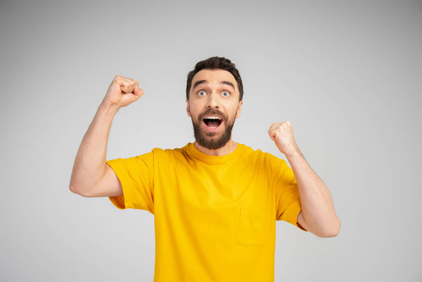 збуджений бородатий чоловік у жовтій футболці кричить і демонструє жест успіху ізольований на сірому
 - Фото, зображення