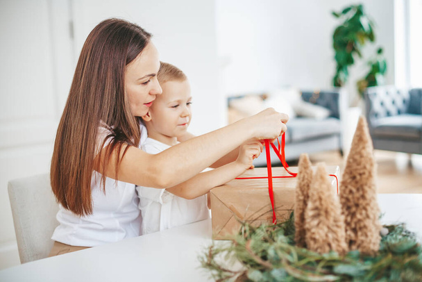 La mamma e il suo piccolo figlio imballano una grande scatola regalo di Natale insieme a casa, legando un fiocco su un grande regalo di Natale. - Foto, immagini