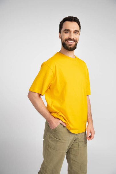 brunette man in yellow t-shirt standing with hand in pocket and smiling at camera isolated on grey - Photo, Image