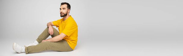 full length of bearded man in beige trousers and yellow t-shirt sitting on grey background, banner - Foto, Imagen