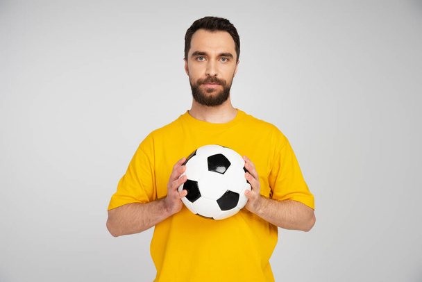 bearded sports fan in yellow t-shirt holding soccer ball while looking at camera  isolated on grey - Zdjęcie, obraz
