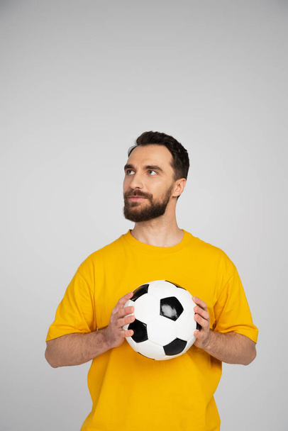 morena barbuda abanico de fútbol en camiseta amarilla sosteniendo pelota y mirando hacia otro lado aislado en gris - Foto, imagen