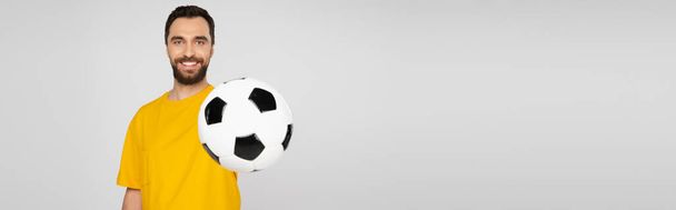 Mutlu ve sakallı spor fanatiği futbol topunu gösteriyor ve gri pankartta izole edilmiş kameraya bakıyor - Fotoğraf, Görsel
