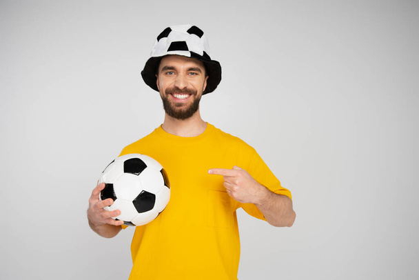 счастливый человек в шляпе футбольного болельщика, указывающий на футбольный мяч, глядя в камеру, изолированную от серого - Фото, изображение