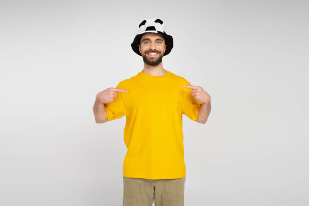 hombre barbudo alegre en el sombrero del ventilador del fútbol y camiseta amarilla que señala con los dedos en sí aislado en gris - Foto, imagen