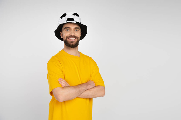 happy bearded man in football fan hat and yellow t-shirt standing with crossed arms isolated on grey - Foto, Imagen