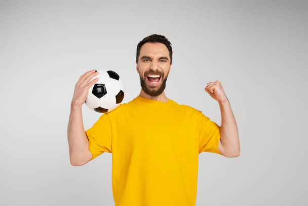 περιχαρής γενειοφόρος άνδρας με μπάλα ποδοσφαίρου δείχνει χειρονομία θριάμβου και ουρλιάζοντας απομονωμένη σε γκρι - Φωτογραφία, εικόνα