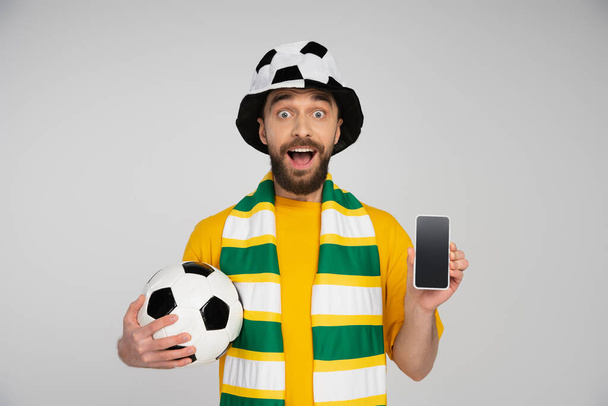 užaslý fotbalový fanoušek drží fotbalový míč a smartphone s prázdnou obrazovkou izolované na šedé - Fotografie, Obrázek