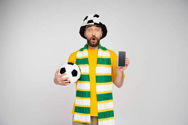 удивленный человек в футбольной шляпе, показывающий смартфон с пустым экраном, изолированным на сером - Фото, изображение