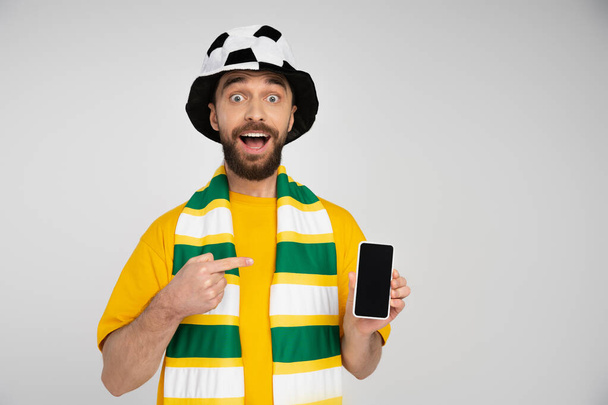 astonished football fan with open mouth looking at camera and pointing at cellphone with blank screen isolated on grey - Foto, immagini