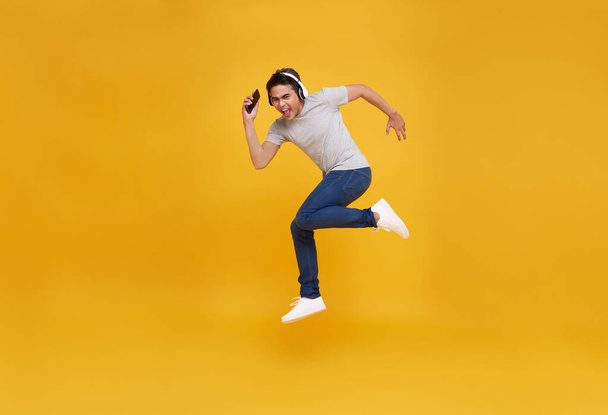 Joven hombre asiático guapo sonriendo sosteniendo teléfono inteligente y saltando usando auriculares inalámbricos escuchando música aislada sobre fondo amarillo. - Foto, Imagen