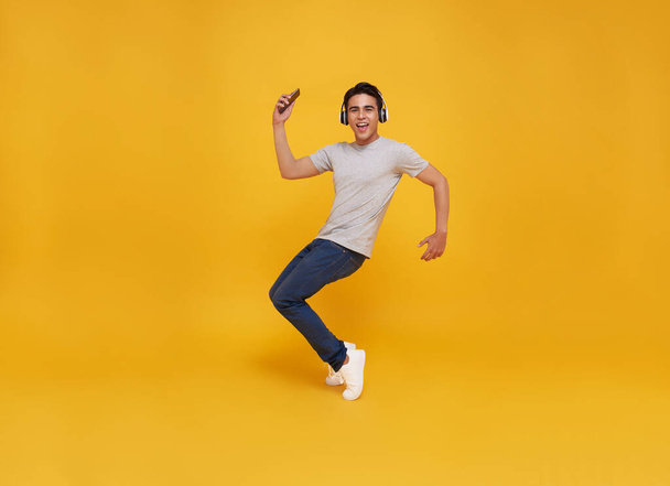 Young handsome Asian man smiling holding smartphone and jumping wearing wireless headphone listening to music isolated over yellow background. - Photo, Image