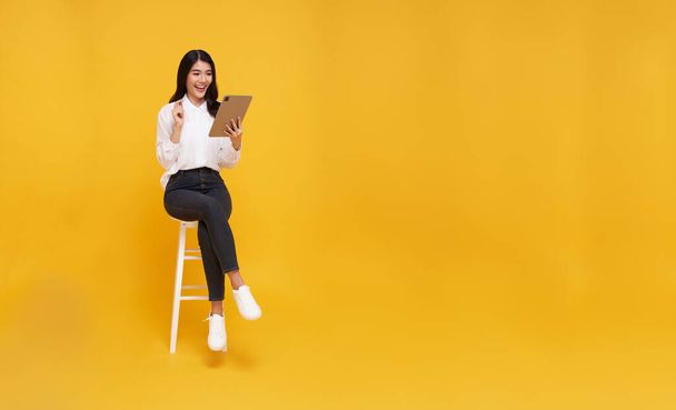 Vrolijke jonge Aziatische glimlach gezicht vrouw met behulp van tablet. Terwijl ze zit op een witte stoel en kijkt op gele kopieerruimte achtergrond. - Foto, afbeelding