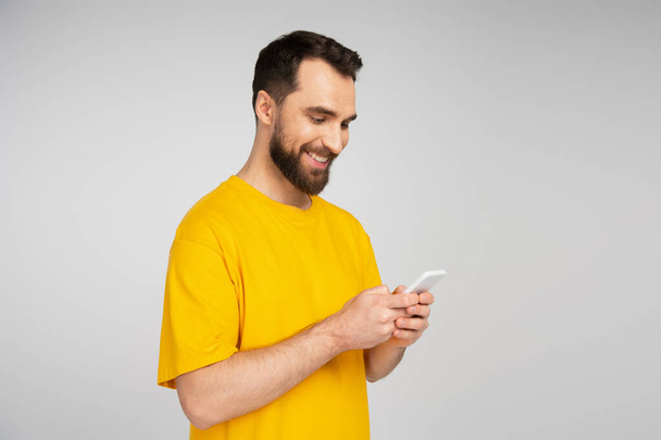 smiling bearded man in yellow t-shirt using mobile phone isolated on grey - Fotografie, Obrázek