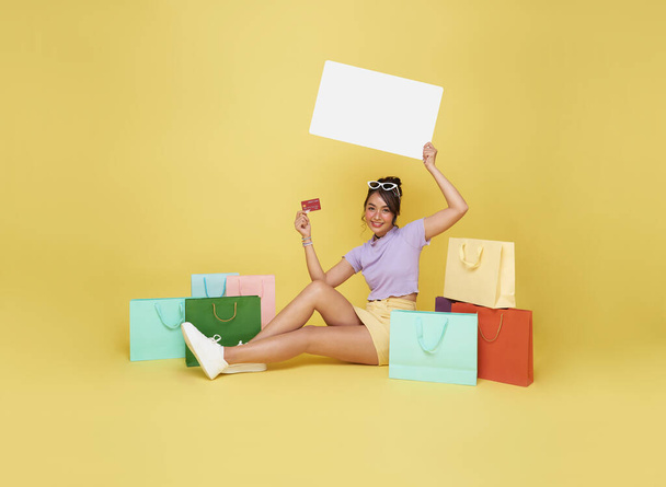 Piękny nastolatek Azji kobieta shopper siedzi z torby na zakupy i trzymając kartę kredytową z pustym billboardem w rękach na żółtym tle. - Zdjęcie, obraz